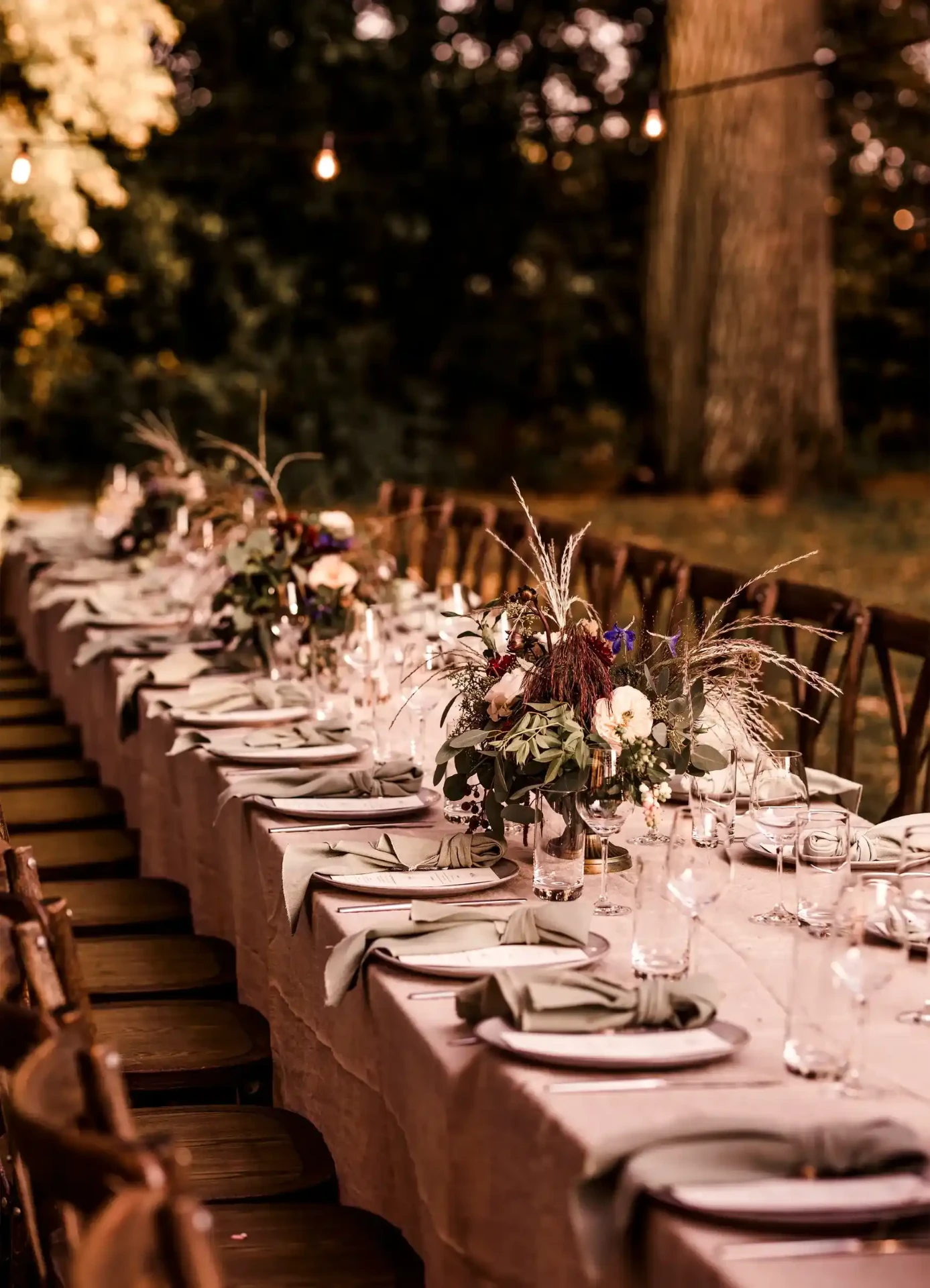 Long wedding table beneath trees with white floral arrangements