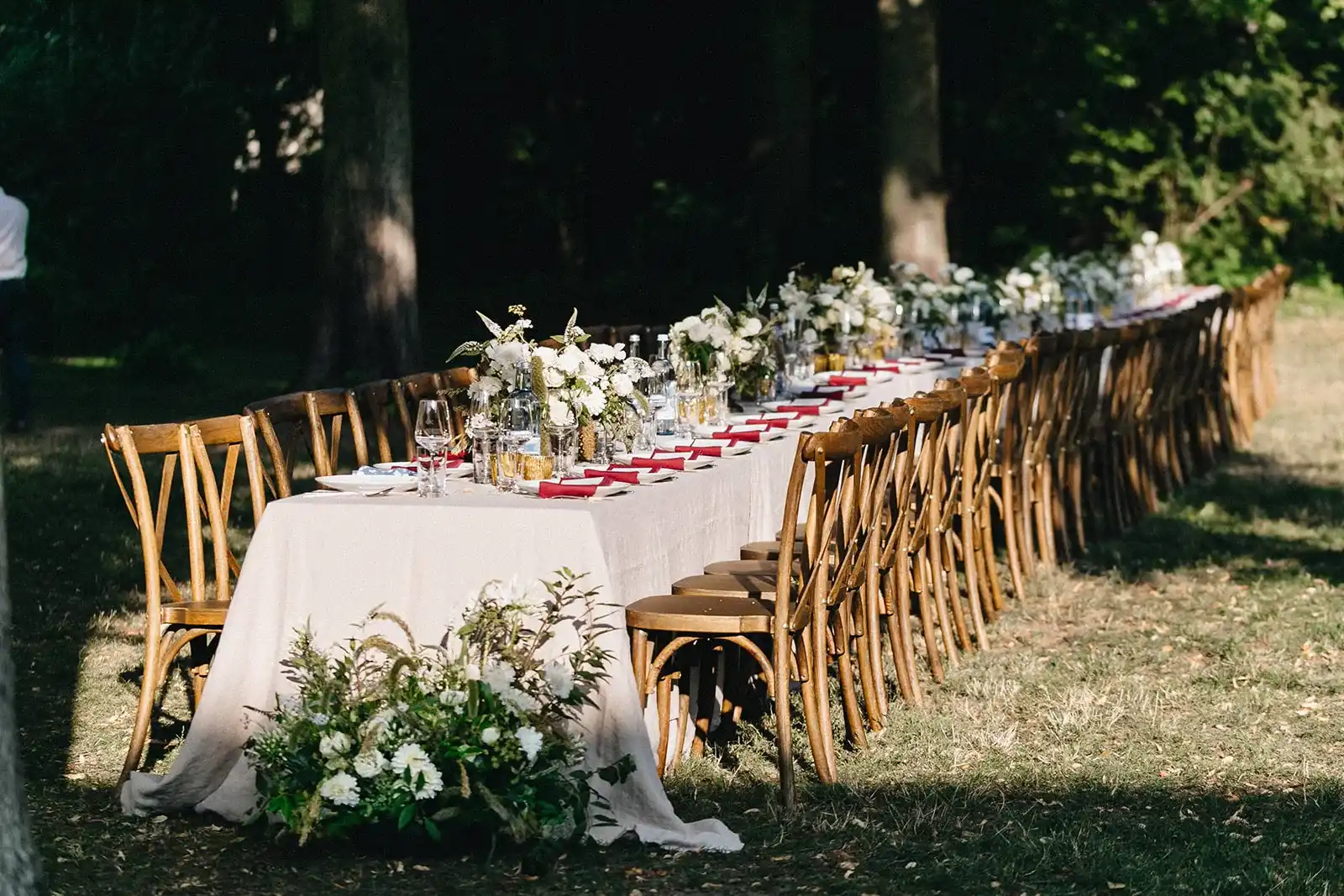 Lange Tafel mit weißen Blumenarrangements und Holzstühlen auf einer Wiese