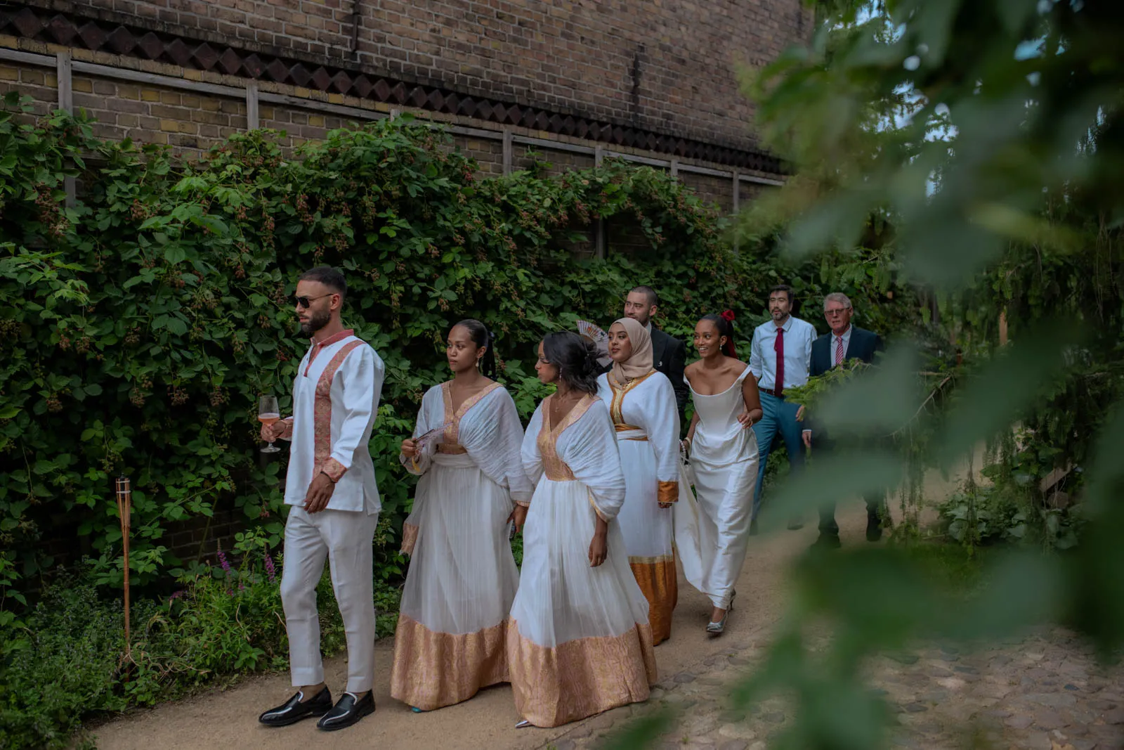 Wedding couple and guests walking beside a brick wall covered in greenery
