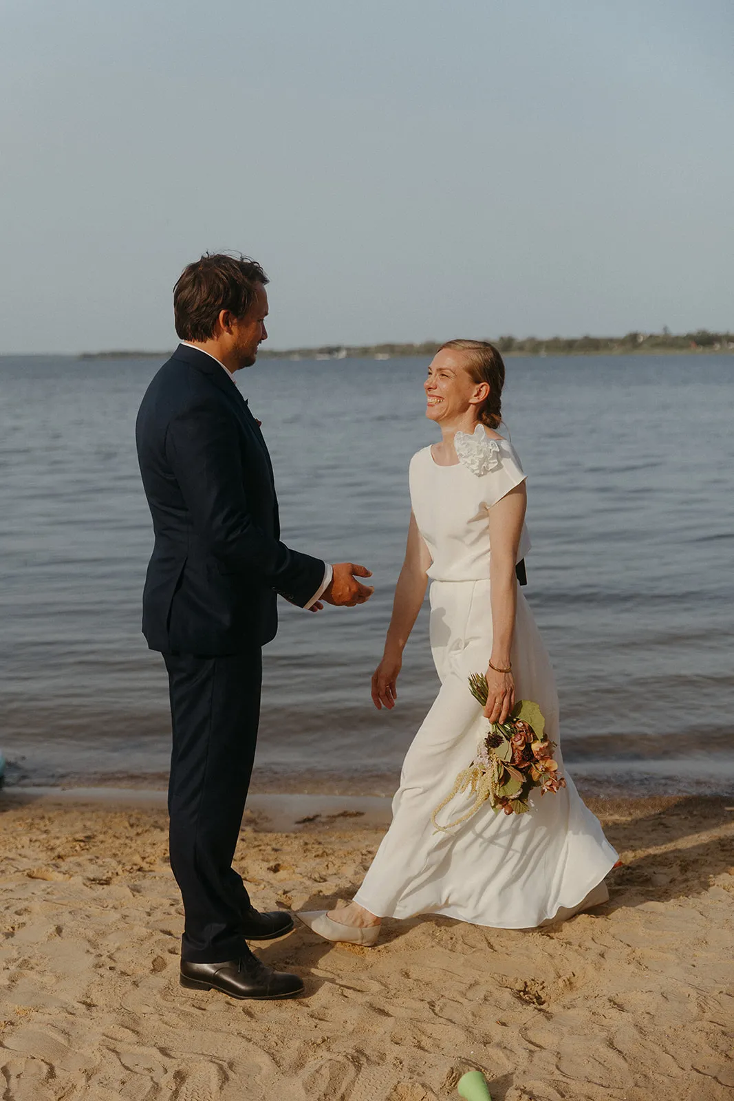 Bride and groom facing each other on the sandy shore of Lake Müritz; the bride holds a bouquet