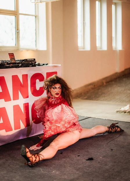 Woman in a pink outfit seated in front of a red “DANCE DANCE” sign