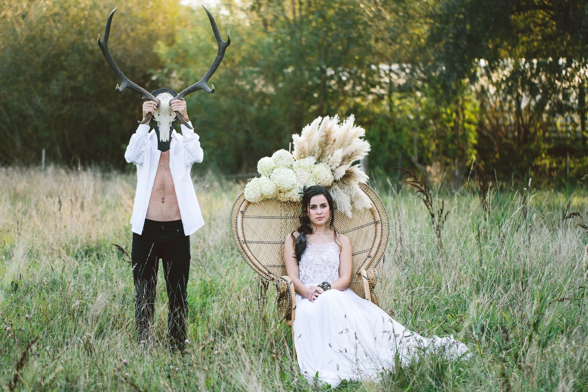 Groom holding a deer skull in front of his face while the bride sits in a rattan chair in tall grass