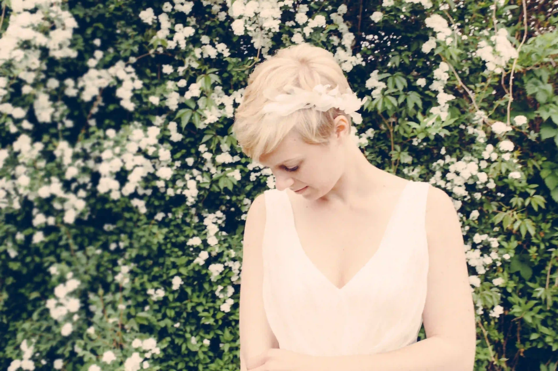 Bride with short blonde hair in a white dress in front of a flowering hedge