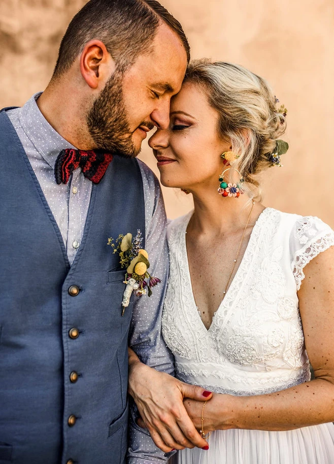 Bride and groom resting their foreheads together; the bride wears a white lace dress and statement earrings