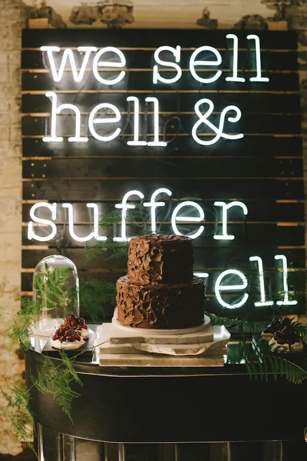 Chocolate cake on marble slabs and cherry pavlova in front of the neon sign “we sell hell & suffer well”