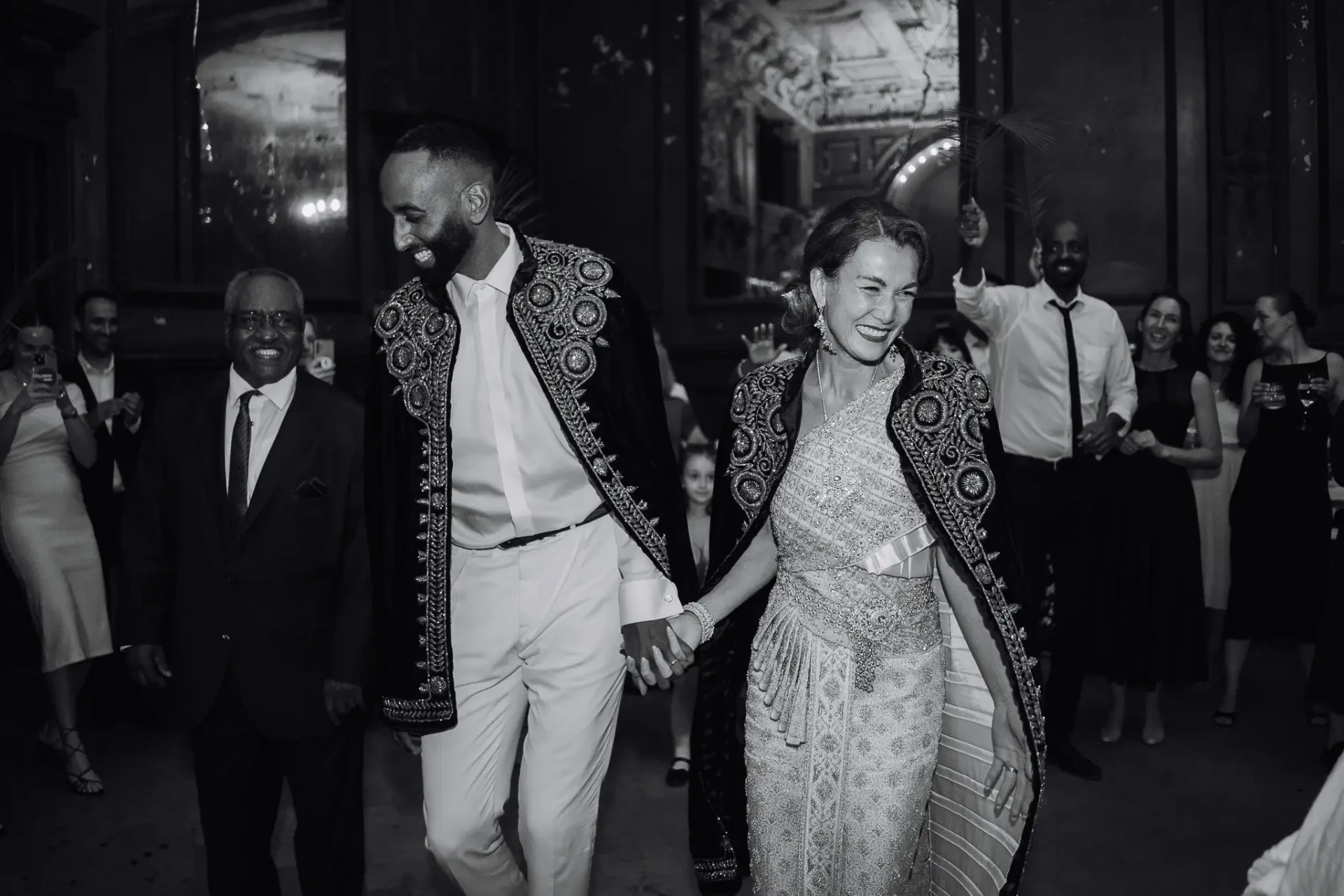Bride and groom in richly embroidered traditional attire walking hand in hand through their wedding guests