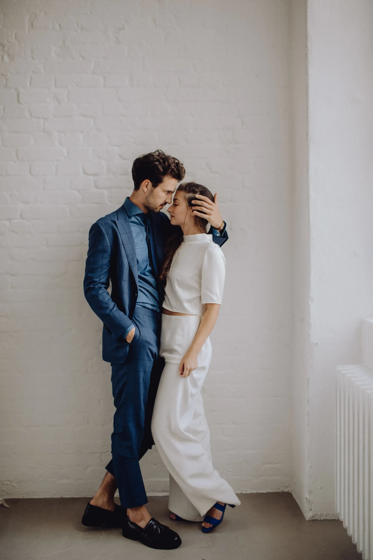 Wedding couple standing against a light wall, the bride wearing a simple white dress