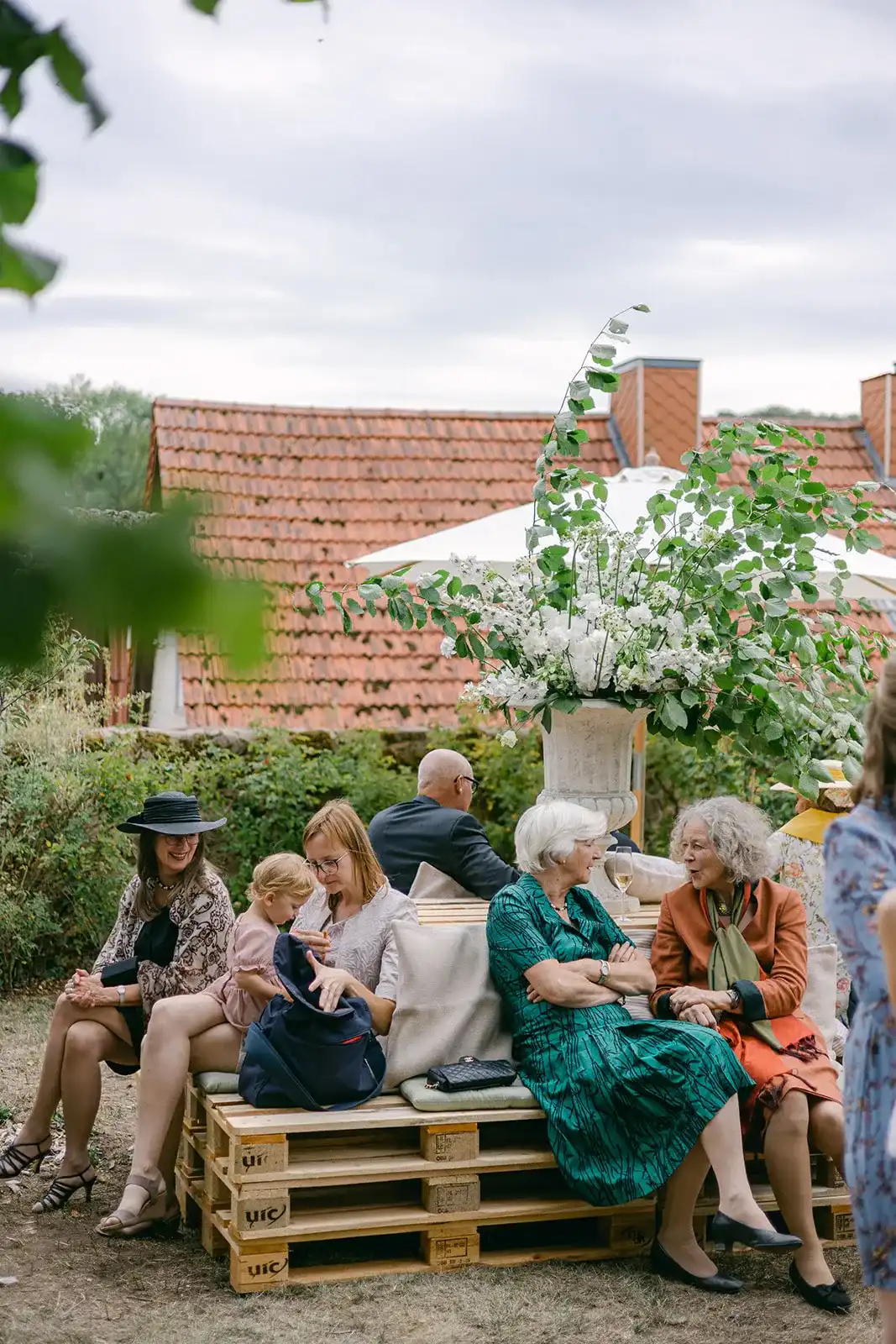 Wedding guests seated on wooden benches in a garden in front of a brick building