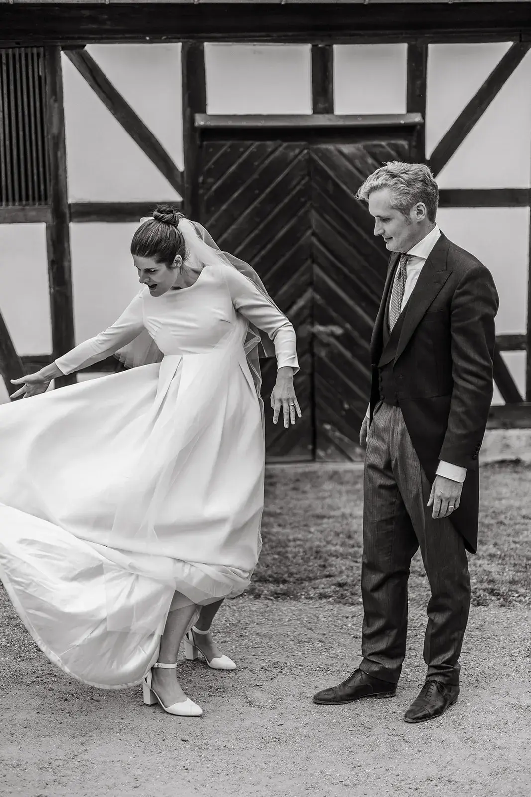 Bride spreading her wedding dress beside the groom in front of a half-timbered building, black-and-white photograph