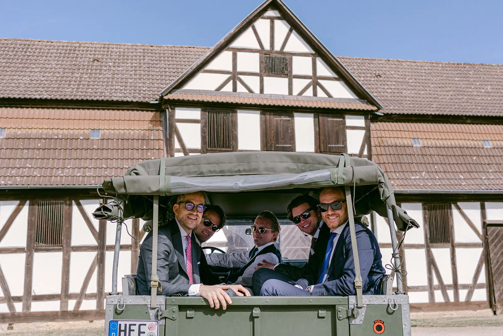 Men in formal attire sitting in a small open vehicle in front of a half-timbered building