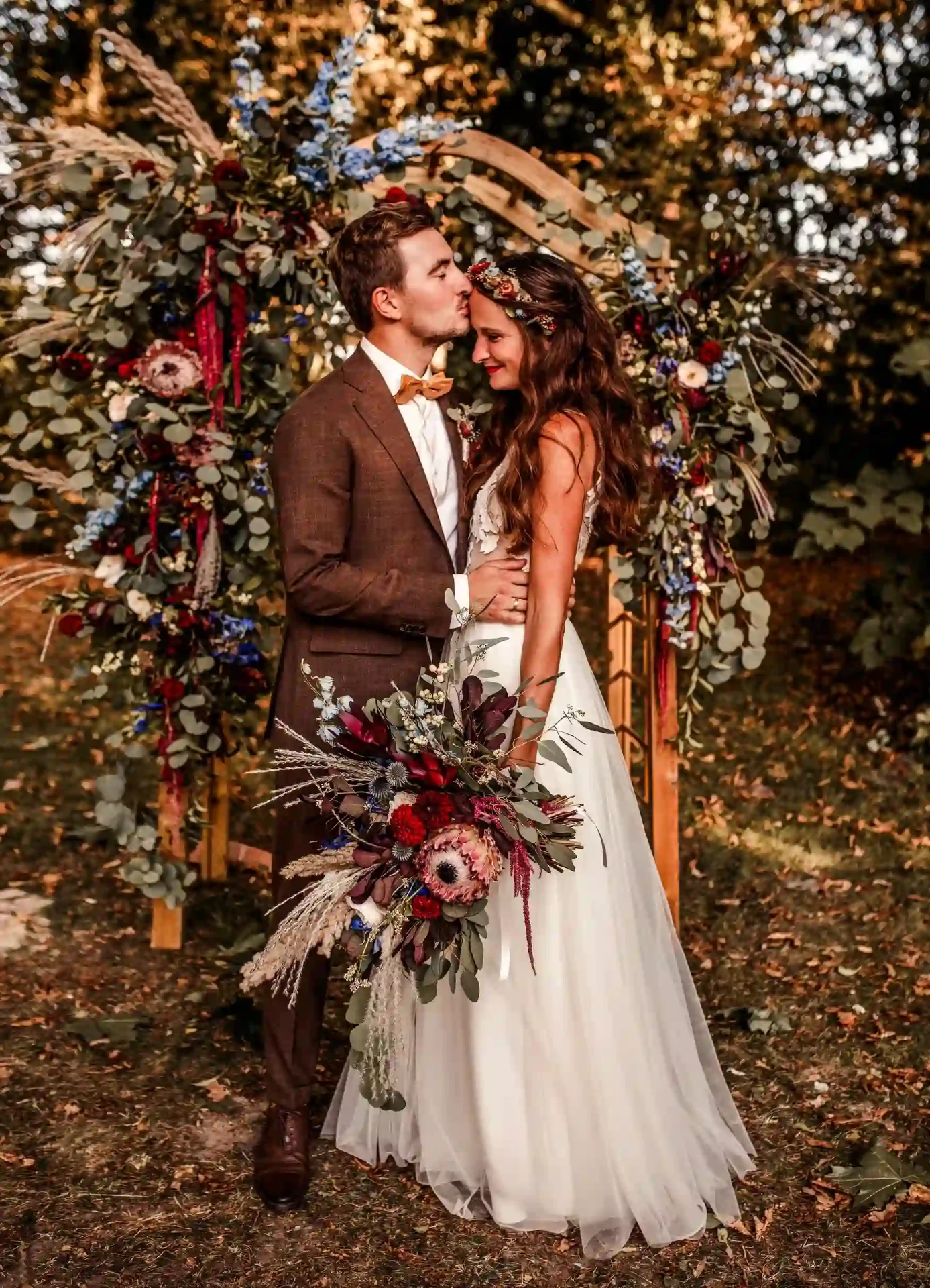 Groom kissing his bride’s forehead beneath a lush floral wedding arch